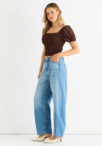 Braunes Smock-Crop-Top mit Puffärmeln, kombiniert mit hellblauen weiten Jeans und cremfarbenen spitzen Absätzen auf einem einfarbigen Hintergrund.
