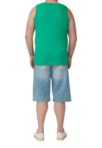 Mężczyzna w zielonej bezrękawnikowej koszulce, jasnoniebieskich szortach dżinsowych i białych trampkach stoi tyłem na białym tle.