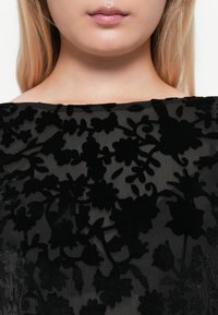 Schwarzes Blumenspitze-Oberteil aus einem transparenten Stoff mit einem erhobenen, samtigen Blumendesign. Der glatte Ausschnitt sitzt direkt über den Schultern.