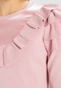 Pull en rose clair avec un col rond, des volants en satin le long des épaules et de la poitrine, et une finition en tissu texturé.