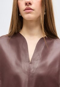 Νεαρή γυναίκα με ίσια ανοιχτό καστανά μαλλιά που φοράει μπορντό δερμάτινο τοπ με V-σχήμα στο ντεκολτέ και ασημένια σκουλαρίκια σταγόνες.