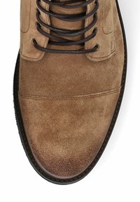 Brązowy zamszowy botek za kostkę z okrągłym noskiem, ciemnobrązowymi sznurowadłami i kontrastującym przeszyciem. Teksturowana powierzchnia oraz elastyczna gumowa podeszwa.