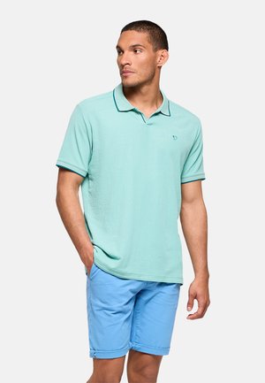 Hombre con el pelo corto, vestido con una camisa polo verde menta y pantalones cortos azul claro, de pie con una mano en el bolsillo, mirando hacia un lado.