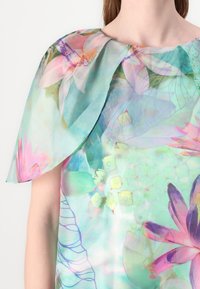 Bloemenprint top met een turquoise achtergrond en roze en groene tinten, semi-doorzichtige stof en een gestructureerd kraagdetail in de vorm van een cape.