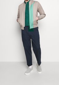 Světlobéžová bomber bunda s zeleným akcentem, zelené polo tričko, tmavě modré volné kalhoty a šedé slip-on boty. Hladká textura, ležérní design.