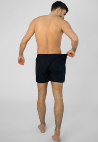 Blauwe zwembroek met een elastische tailleband, zichtbare stiksels en een snit tot halverwege de bovenbeen, gedragen door een persoon die met de rug naar de kijker staat.