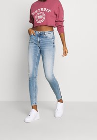 Jeans ajustados de color azul claro con parches descoloridos, combinados con una sudadera corta de color burdeos con un estampado gráfico blanco. Zapatillas blancas complementan el look.