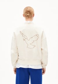 Crème-kleurige sweatshirt met een grote bruine lijntekening van een duif op de rug. Ribgebreide mouwen en zoom, losse pasvorm, gedragen over een wit shirt.