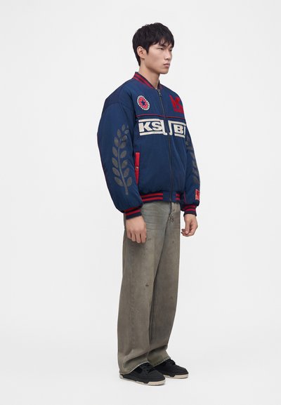 Jeune homme debout portant une veste letterman bleue et rouge avec des logos, un pantalon gris ample et des baskets noires sur un fond blanc uni.