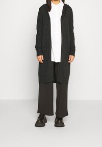 Tmavě šedý pletený kardigan s dlouhými rukávy, předními kapsami a kapucí, nošený přes bílou košili na knoflíky a tmavé kalhoty s širokými nohavicemi.