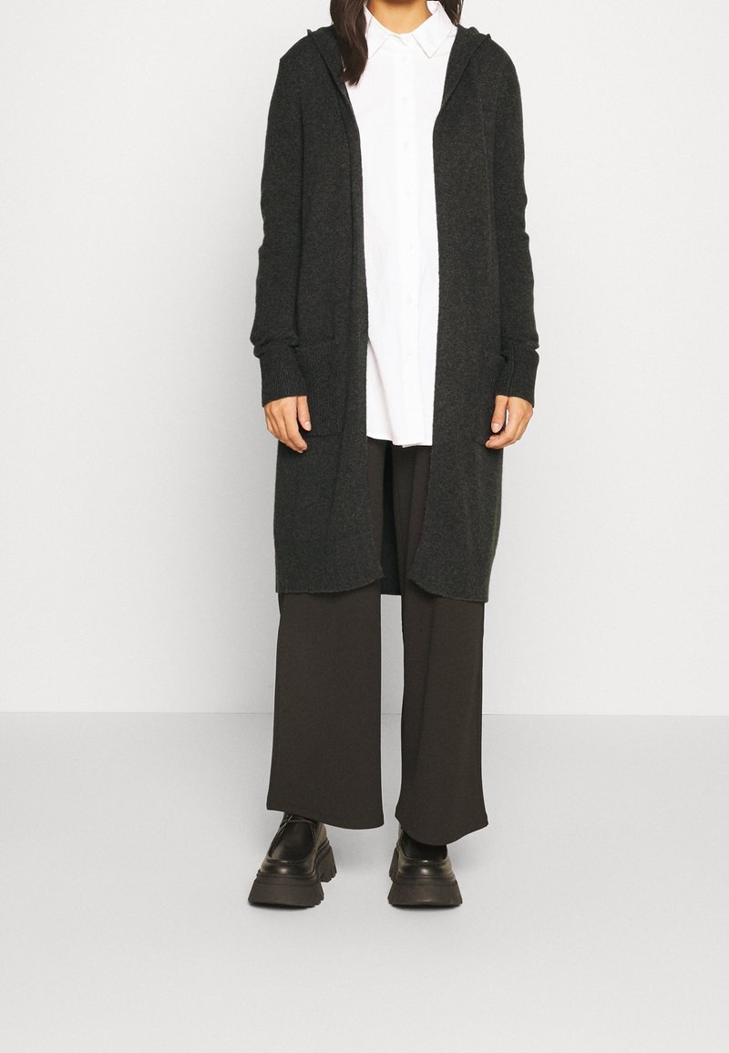 Tmavě šedý pletený kardigan s dlouhými rukávy, předními kapsami a kapucí, nošený přes bílou košili na knoflíky a tmavé kalhoty s širokými nohavicemi.