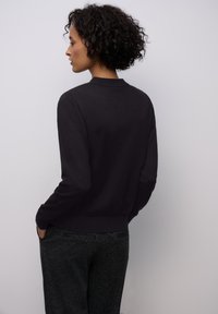 Černý kardigan s dlouhým rukávem a žebrovaným lemem a límcem, vyrobený z měkkého, hladkého materiálu; v kombinaci s tmavými vzorovanými kalhotami.