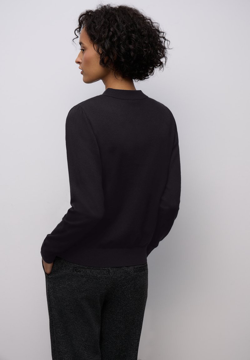 Černý kardigan s dlouhým rukávem a žebrovaným lemem a límcem, vyrobený z měkkého, hladkého materiálu; v kombinaci s tmavými vzorovanými kalhotami.