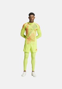 Felle groene sportoutfit met strakzittende lange mouwen en shorts met oranje geometrische patronen. Schoeisel in het wit, met een slank ontwerp.