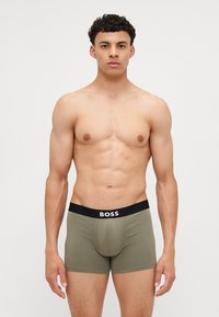Maslinasto zeleni bokserice s crnim elastičnim pojasom na kojem je bijeli logo "BOSS". Glatka, rastezljiva tkanina s prilagođenim dizajnom.