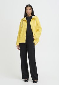 Veste imperméable jaune avec une capuche blanche, devant à boutons et cordons de serrage, portée par-dessus un haut noir et un pantalon noir ample.