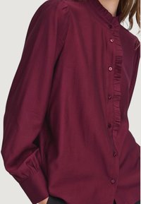 Blusa bordeaux a maniche lunghe con alto colletto, dettaglio arricciato frontale e bottoni perlescenti. Realizzata in un morbido tessuto testurizzato.