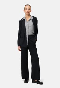 Czarna, dopasowana marynarka na czarno-białej paskowanej koszuli, zestawiona z szerokimi czarnymi spodniami i czarnymi mokasynami; gładkie tekstury tkanin.