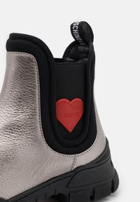 Botte gris métallisé avec panneau latéral élastique noir, patch rouge en forme de cœur portant l'inscription « LOVE MOSCHINO » et détail de semelle noire épaisse.