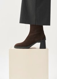Brown suede ankle boot with a rounded toe and a chunky black heel, set on a light wooden block surface against a plain backdrop.