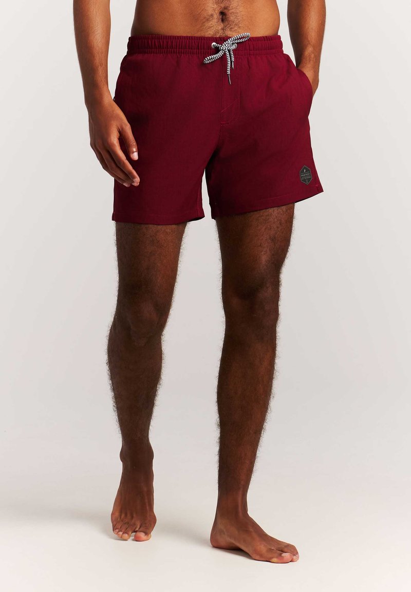 Mann mit dunkler Haut, der burgunderrote Badeshorts mit weißem Kordelzug trägt, steht barfuß vor einem einfarbig hellen Hintergrund.