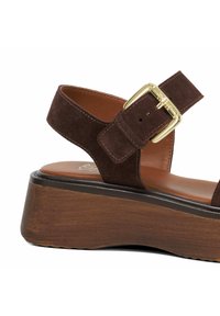 Brown suede sandal with a wide strap, gold buckle detail, and a thick wooden wedge heel. Smooth tan insole with stitching accents.