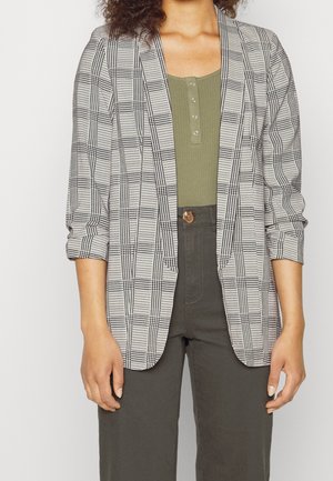 Mujer con blazer de cuadros en blanco y negro sobre una blusa verde oliva con botones y pantalones de talle alto de color gris oscuro, de pie frente a un fondo blanco.
