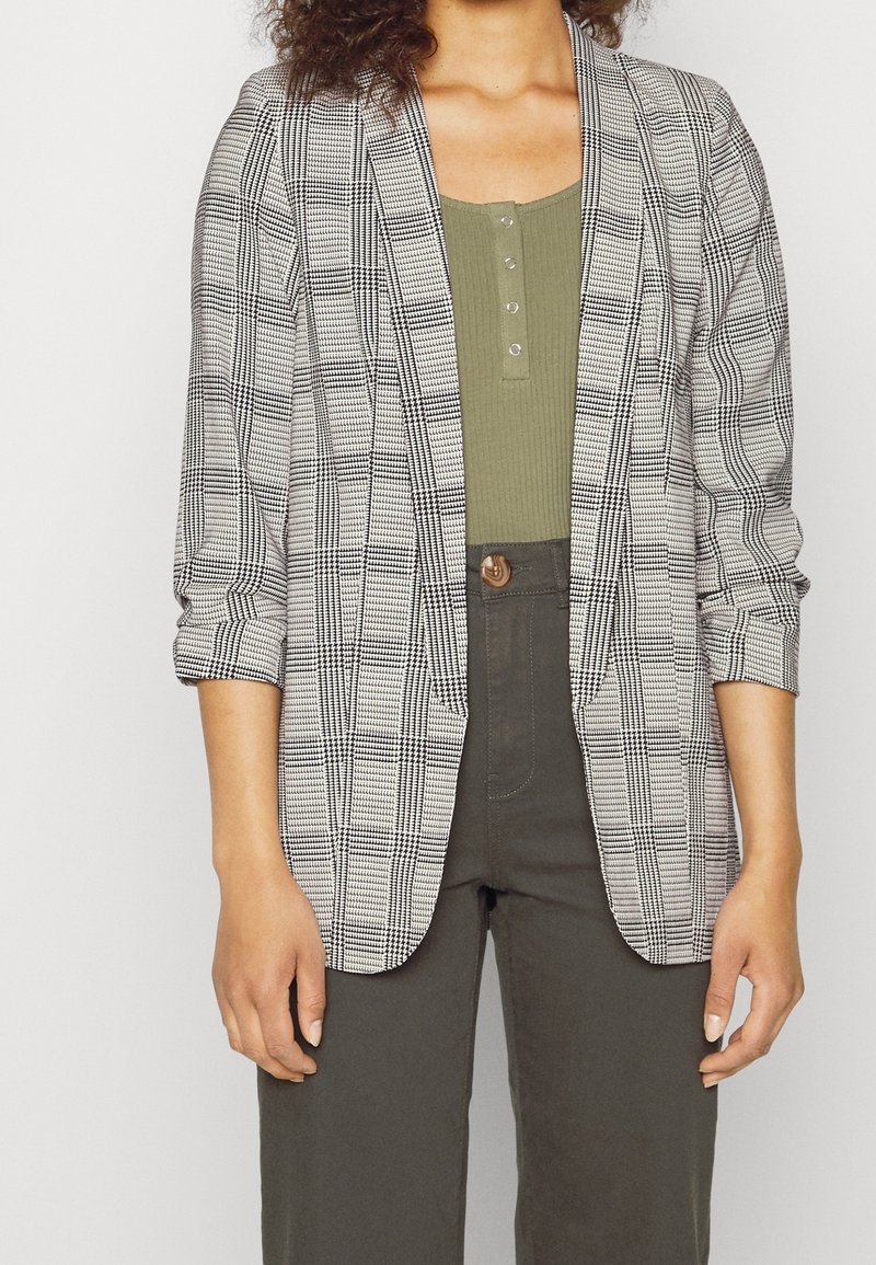 Femme portant un blazer à carreaux noir et blanc sur un haut vert olive boutonné et un pantalon taille haute gris foncé, debout sur un fond blanc.