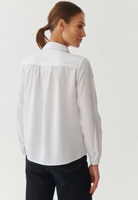 White long-sleeve shirt featuring a back yoke seam, soft fabric, and a curved hem. Paired with dark jeans, highlighting a casual style.