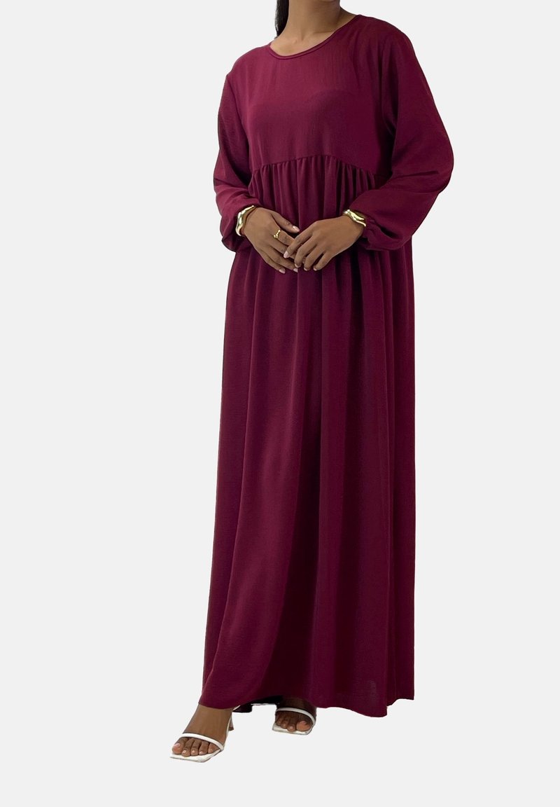 Femme portant une robe bordeaux à manches longues, longue jusqu’au sol, avec une taille froncée, des talons blancs ouverts à l’avant et des bracelets dorés aux deux poignets.