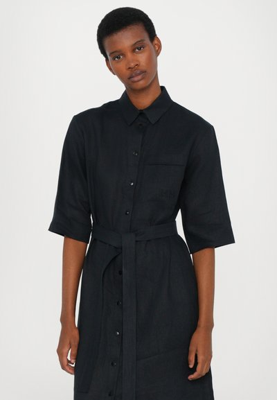 Vestido de camisa em linho preto com fecho de botões na frente, mangas curtas e cinto na cintura. Possui um bolso no lado esquerdo e uma textura subtil.
