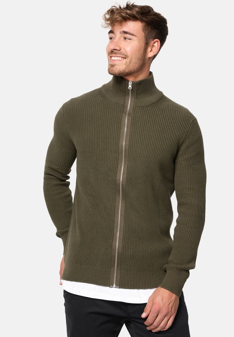 Suéter de cremallera de color verde oliva con canalé y cuello alto, que presenta una cremallera de metal, mangas largas y un diseño ajustado.