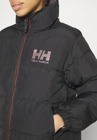 Personne portant une doudoune noire Helly Hansen avec la main dans la poche et le col montant partiellement fermé par une fermeture éclair.