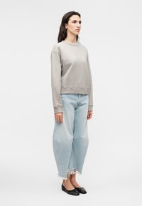 Mujer de pie con sudadera gris de manga larga, vaqueros anchos de pierna con dobladillo deshilachado en azul claro y bailarinas negras sobre fondo blanco.