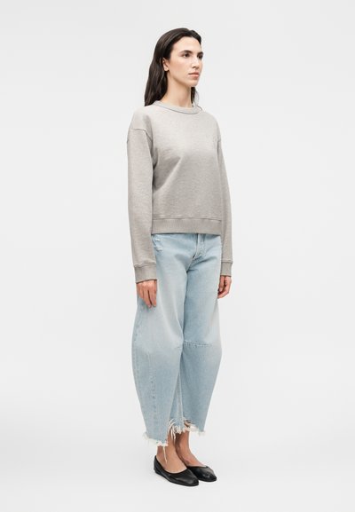 Γυναίκα στέκεται φορώντας γκρι μακρυμάνικο φούτερ, ανοιχτό μπλε φαρδιά ξεφτισμένα τζιν και μαύρα μπαλαρίνες σε λευκό φόντο.