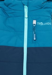 Mėlyna ir tamsiai mėlyna pūkinė striukė su šviesiai mėlynu užtrauktuku ir "TROLLKIDS" logotipu virš užtraukiamos krūtinės kišenės.