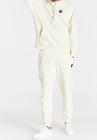 Person bär en krämfärgad hette och matchande sweatpants med svarta broderade detaljer, tillsammans med vita sneakers, medan hen står framför en vit bakgrund.