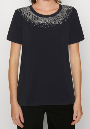 Chemise noire à manches courtes avec encolure ronde, ornée de clous argentés formant un motif décoratif le long du col.