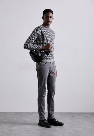 Pull en motif noir et blanc, jeans texturés gris, mocassins noirs et sac à bandoulière noir. Le modèle se tient devant un fond neutre.