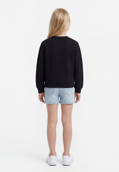 Copil cu păr blond lung, stând cu fața spre spate, purtând un hanorac negru, pantaloni scurți de blugi albastru deschis și adidași albi.