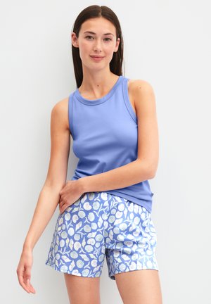 Jeune femme portant un haut lavande sans manches et un short bleu avec un motif de coquillages blancs, debout devant un fond blanc.