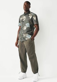 Camisa de manga corta, de color verde oscuro con estampado floral en tonos azul claro y beige, combinada con pantalones tapered color oliva claro y zapatillas blancas.