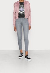 Chaqueta bomber de satén rosa con puños de canalé, camiseta gráfica negra, jeans ajustados grises y zapatillas Vans negras con detalles en blanco.