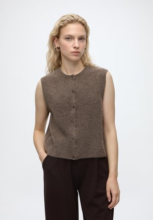 Femme blonde portant des boucles d’oreilles créoles en or, un gilet marron sans manches boutonné et un pantalon marron foncé, debout avec une main dans la poche.