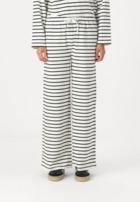 White fabric pants with black horizontal stripes, featuring an elastic waistband with a drawstring. Loose fitting with wide legs.