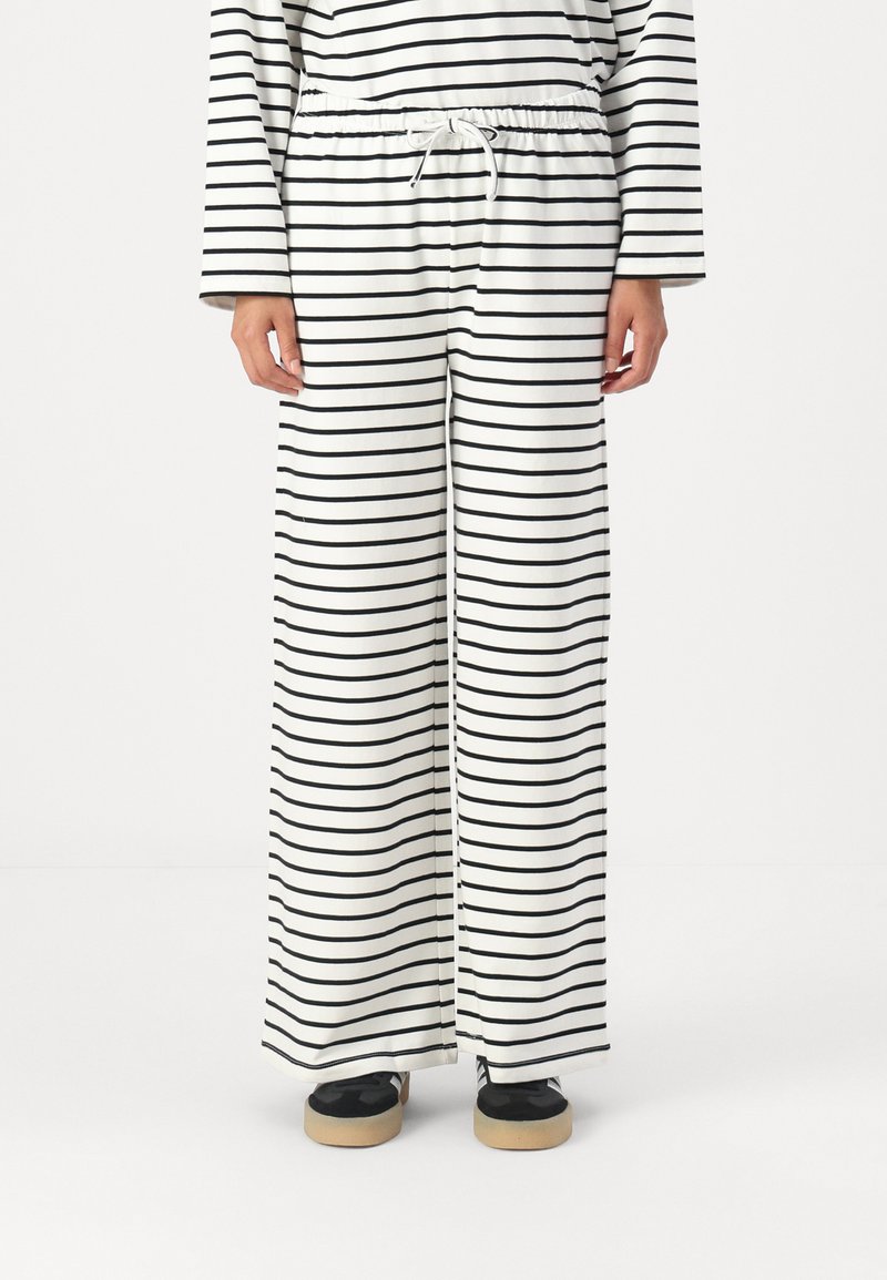 White fabric pants with black horizontal stripes, featuring an elastic waistband with a drawstring. Loose fitting with wide legs.