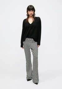 Schwarzer Strickpullover mit V-Ausschnitt und offenem Strickmuster, kombiniert mit weit geschnittenen Hosen mit einem schwarz-weißen geometrischen Muster.