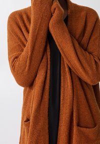 Cardigan couleur rouille en tissu doux et texturé. Il présente une coupe ample avec un devant ouvert et deux poches avant.