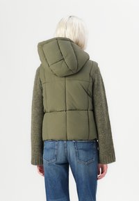 Maslinasto zeleni puffer prsluk s opaženim dizajnom, kapuljačom i teksturiranim sivim rukavima. Uparen s plavim trapericama.