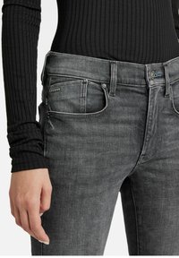 Vaqueros de mezclilla gris oscuro con corte ajustado, que cuentan con cinco bolsillos, costuras sutiles y una etiqueta de marca en el bolsillo frontal.